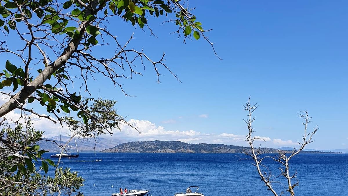 Agni Bay in Corfu, seen from the hillside through olive branches, with motorboats anchored in turquoise water and the mainland mountains beyond