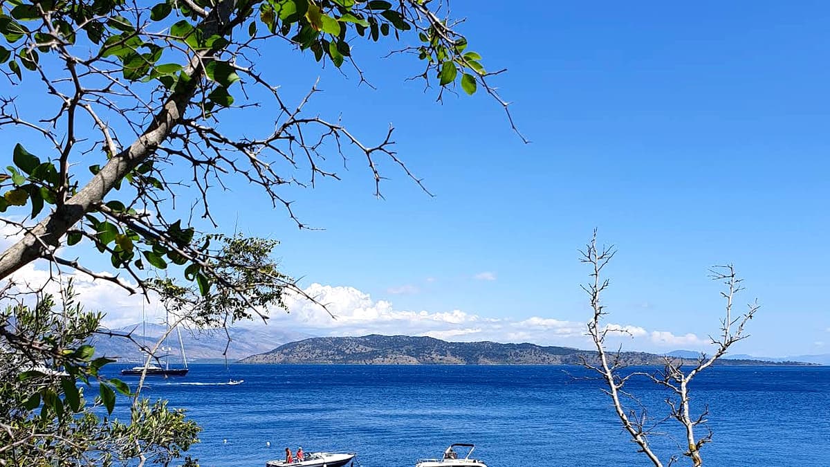Agni Bay in Corfu, seen from the hillside through olive branches, with motorboats anchored in turquoise water and the mainland mountains beyond