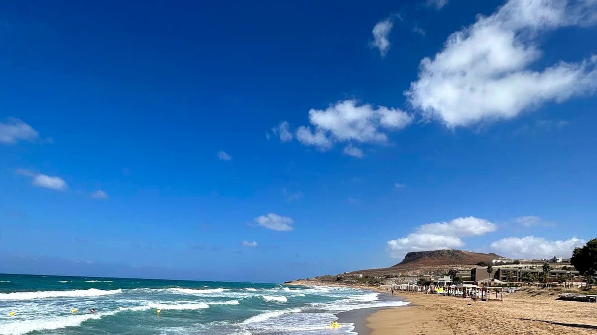 Amnissos Beach in Heraklion, Crete: long curving golden sand with waves on the left and the Paliochora hill rising at the east end of the bay