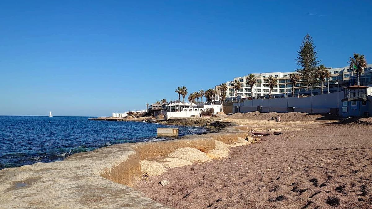 Buġibba Perched Beach: the terrace of imported sand raised above the rocky shoreline of St Paul's Bay, with palm trees and a white hotel block behind and the rocky point beyond
