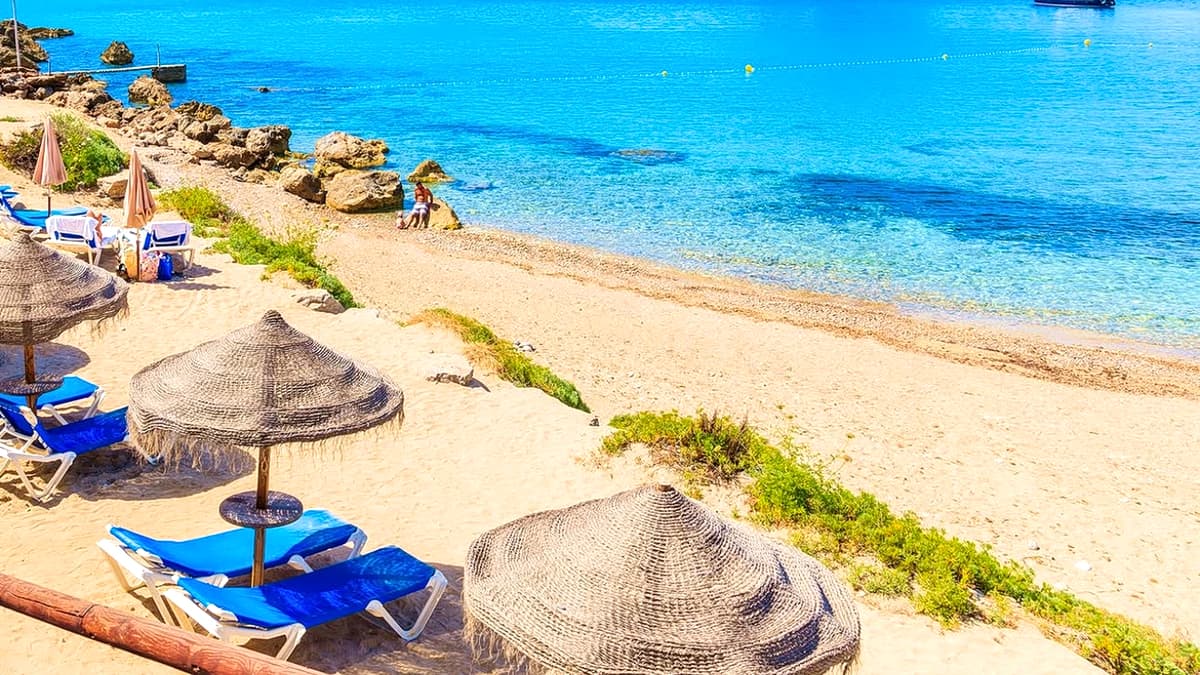 Cala d'Hort beach with straw parasols and blue sun loungers on the sand, turquoise water, and the rock of Es Vedrà with Es Vedranell rising from the sea offshore
