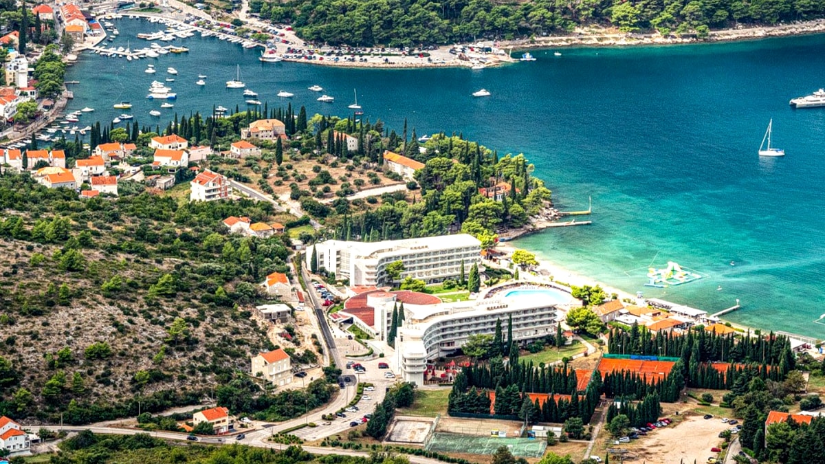 Aerial view of the Cavtat peninsula south of Dubrovnik: a small old town with terracotta roofs, two sheltered bays full of moored sailboats, and pine-forested coastline running out to the open Adriatic