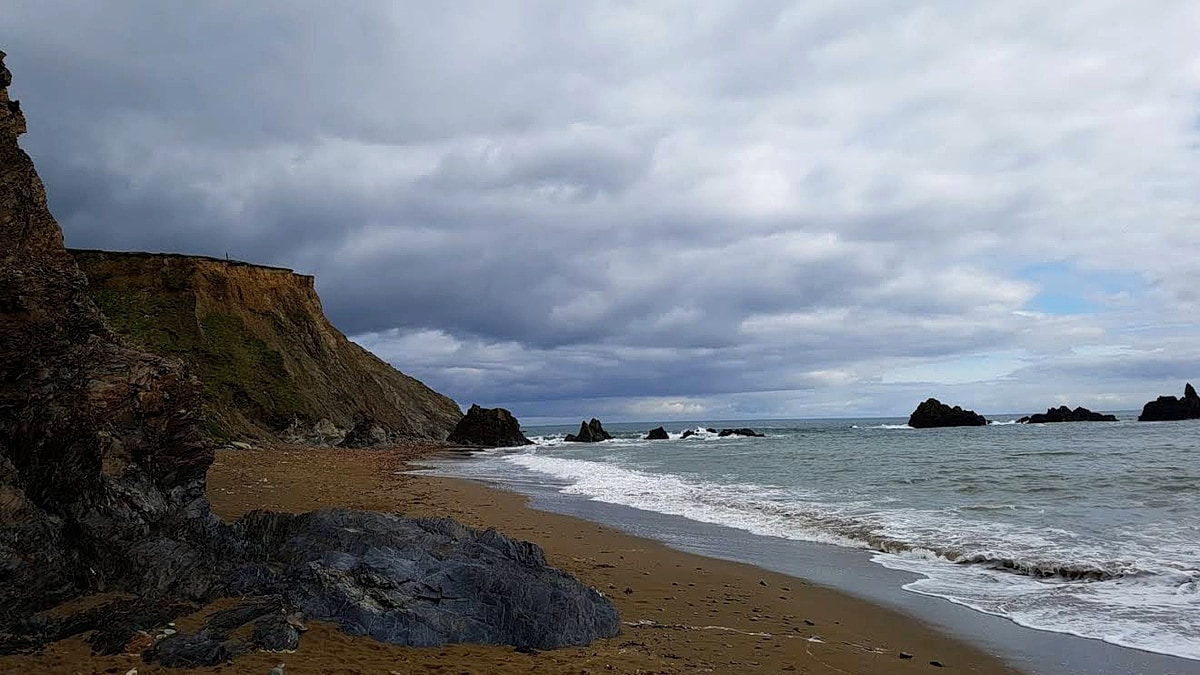 Kilfarrasy Beach on Ireland's Copper Coast under a heavy grey sky: a small cove of tan Atlantic sand below a low headland cliff to the left, a dark rock outcrop on the beach, Atlantic surf breaking on the shore, and rocky sea stacks standing offshore