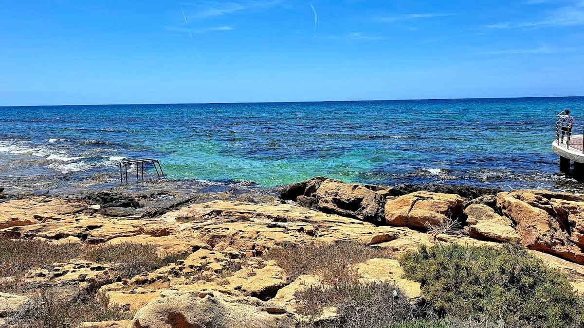 Loukkos Tou Mandi Beach in Ayia Napa, Cyprus: a rocky cove with the steel ladder pier on the left and a wooden viewing platform on the right, looking out at the south-Cyprus turquoise water