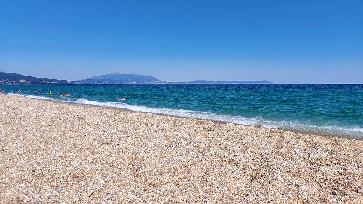 View along Nea Vrasna Beach on the Strymonian Gulf: a broad shelf of pale coarse sand and fine pebble in the foreground, calm blue water reaching out to a long mountain range on the far side of the gulf, a few swimmers in the shallows under a clear deep-blue sky