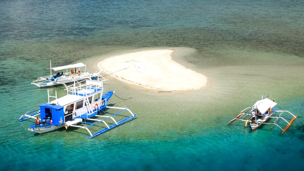 Aerial view of a sandbar with two bangka outrigger boats moored in clear turquoise water off Palawan, with forested headlands behind