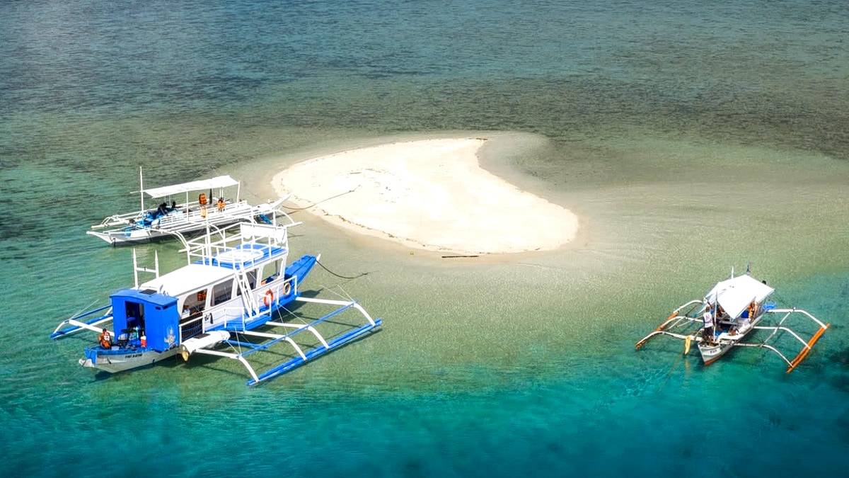 Aerial view of a sandbar with two bangka outrigger boats moored in clear turquoise water off Palawan, with forested headlands behind