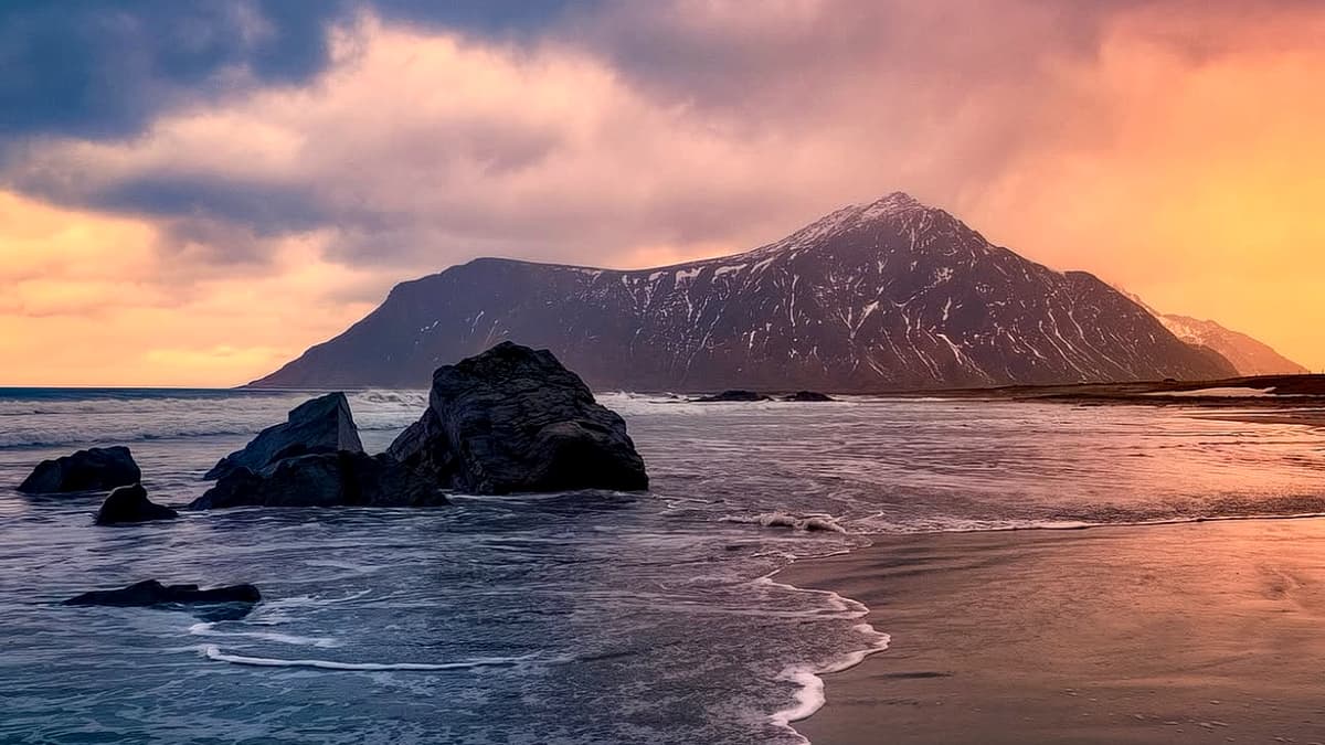 Skagsanden Beach at dusk: a wave sliding over the wet sand flats in a line of white foam, dark rocks on the shore and the snow-streaked ridge of the headland against an orange evening sky