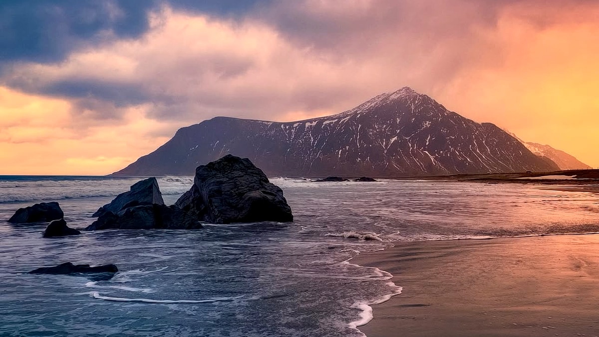 Skagsanden Beach at dusk: a wave sliding over the wet sand flats in a line of white foam, dark rocks on the shore and the snow-streaked ridge of the headland against an orange evening sky